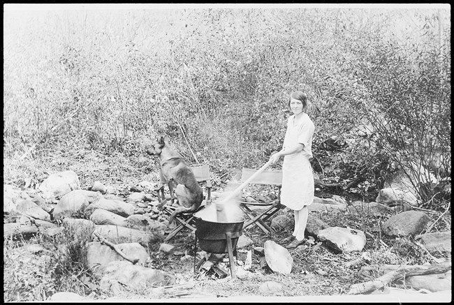 Missie Oakley making apple butter, 1933 A woman uses a long stick to stir a steaming pot over an outdoor fire. A dog sits beside her on one of two camp chairs.