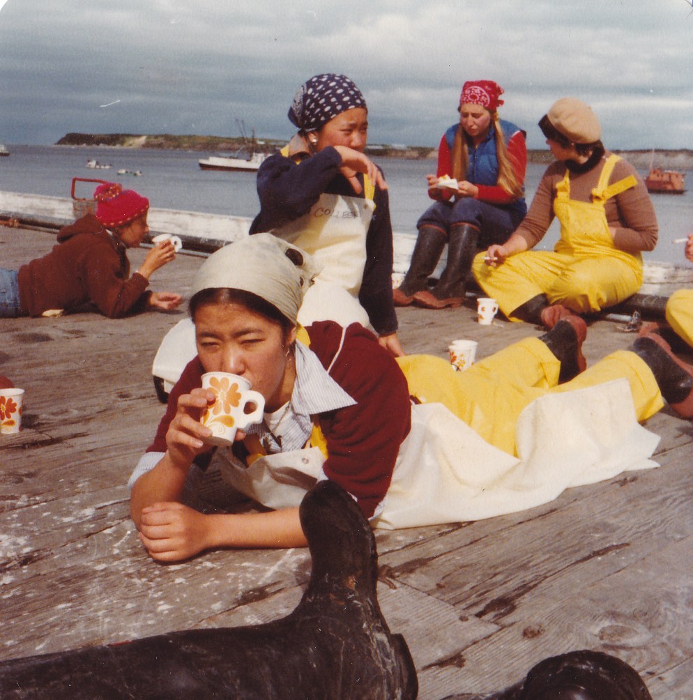 A group of cannery workers, wearing bandannas, boots, and overalls, hold mugs as they sit and lounge on a wooden dock