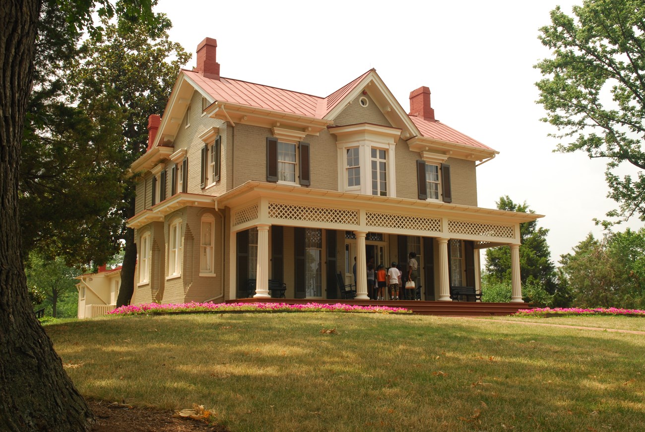 Low flower beds, mature trees, and lawn surround a two-story house with a wide porch.