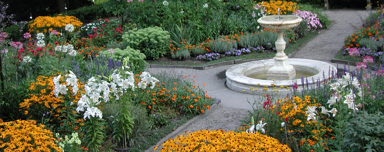 MABI Fountain Terrace Designed Landscape A round fountain on a pedestal stands where two paths cross in a garden of colorful flowershs cross in a garden