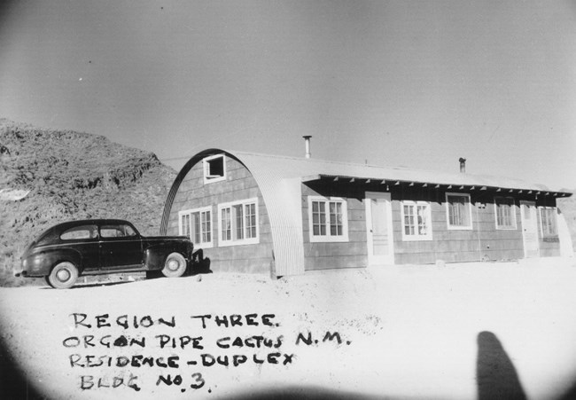A 1950s car is parked beside a quonset hut, shaped like a long half-cylinder with windows and doors.