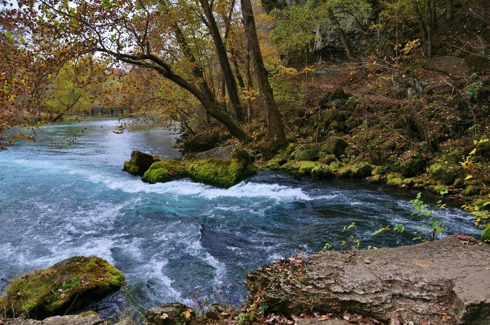 Big Spring causes white-tipped movement on the water, surrounded by mossy rocks and fall foliage