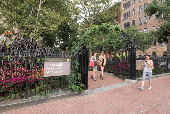 Plants and flowers grow around the metal fence around Christopher Park, where a sign says "Stonewall National Monument"