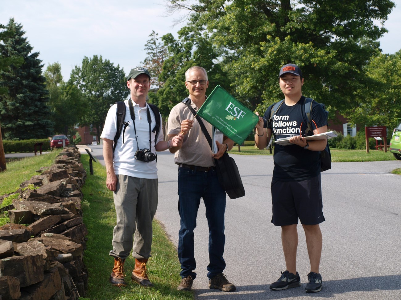 Three men, one with a camera and one with an "ESF" flag, beside a low stone wall.