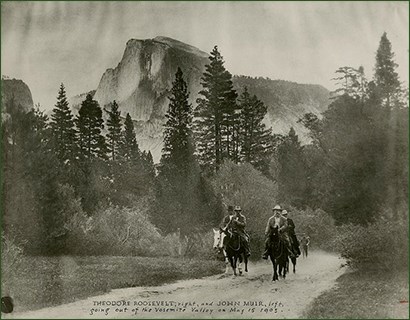 lt= Historic image of four men on horses riding a trail, mountains and trees in the background.