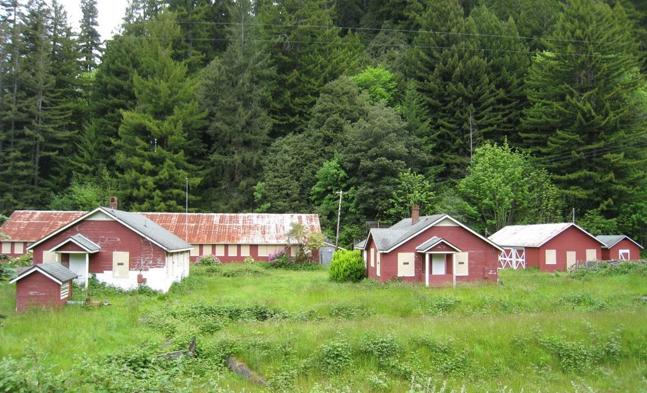 PCFH landscape One-story wooden buildings, red with white trim, are clustered in a level grassy area with a tree-covered hillside in the background