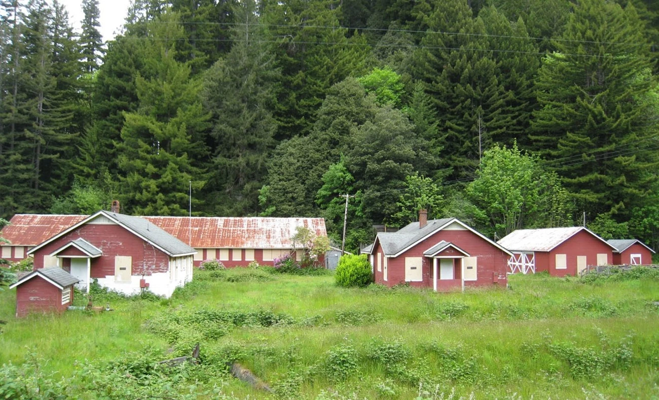 PCFH landscape One-story wooden buildings, red with white trim, are clustered in a level grassy area with a tree-covered hillside in the background