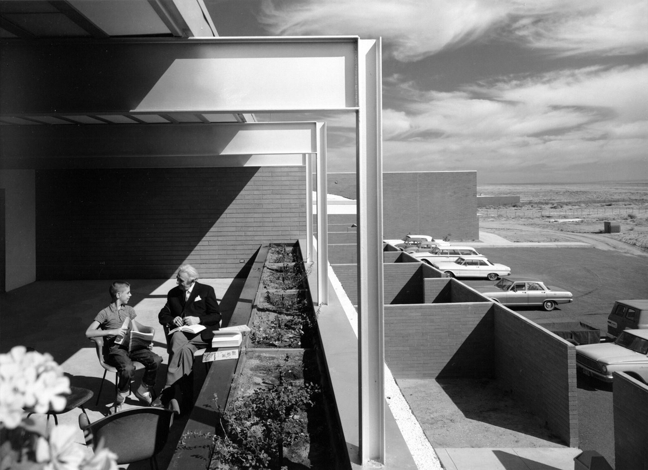 A man and a boy sit in chairs on an apartment balcony at the Painted Desert Community Complex, overlooking brick-walled courtyards and a parking lot