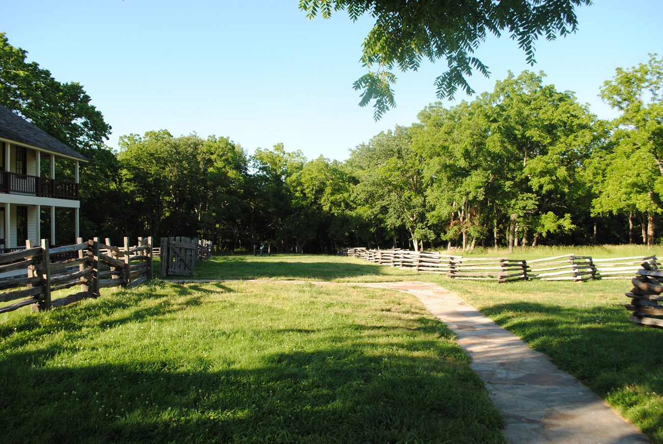 Wooden fences along a path through an open area of grass, with a two-story structure to the left and a border of leafy trees.