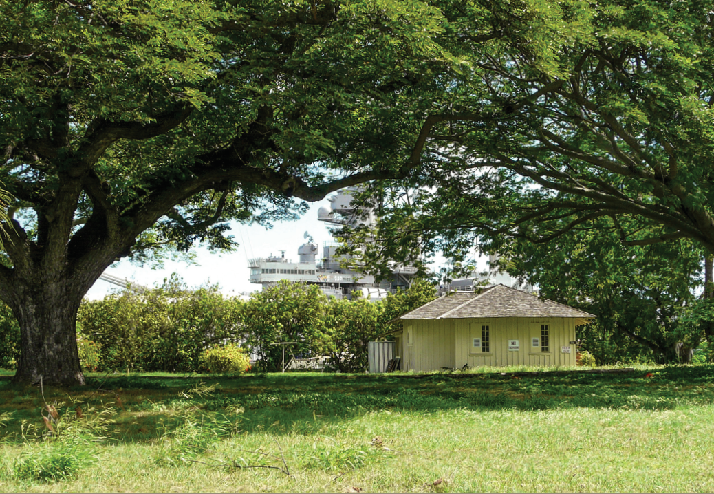 CPO Bungalow 90 A one-story wood-frame structure stands near a wall of vegetation, beyond a lawn shaded by tall leafy trees. A battleship is partly visible in the background, through the trees.