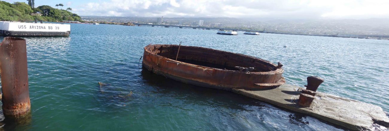View from the USS Arizona Memorial A view over greenish-blue water includes a round, metal section of the USS Arizona protruding from the water's surface. A series of mooring quays and distant land are visible in the background.