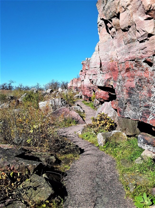 Pink Sioux Quartzite cliffs under a clear, blue sky with a paved trail running between the cliffs and the tallgrass prairie next to them