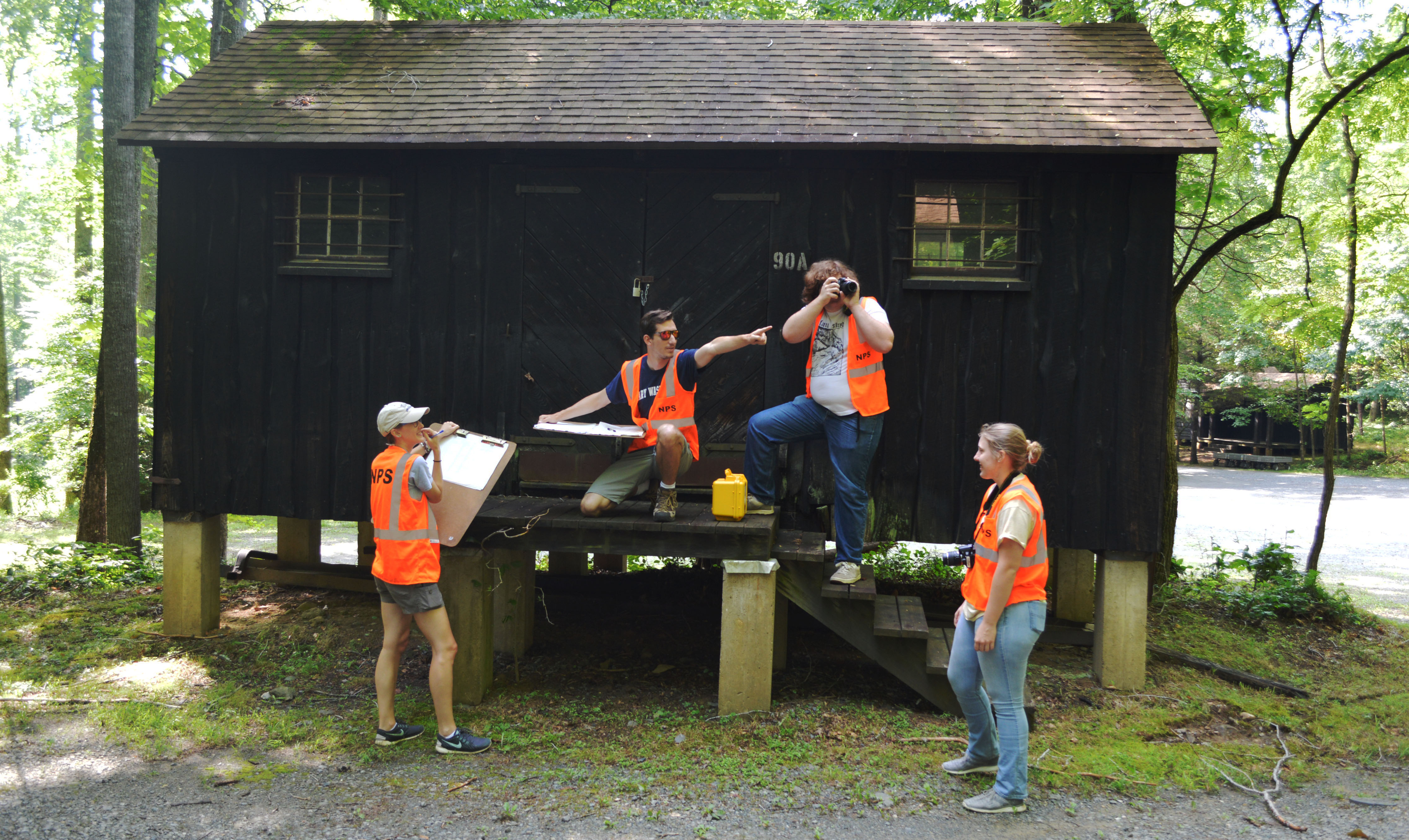 Two people stand on the porch and two stand on the ground in front as they document a small wooden camp cabin