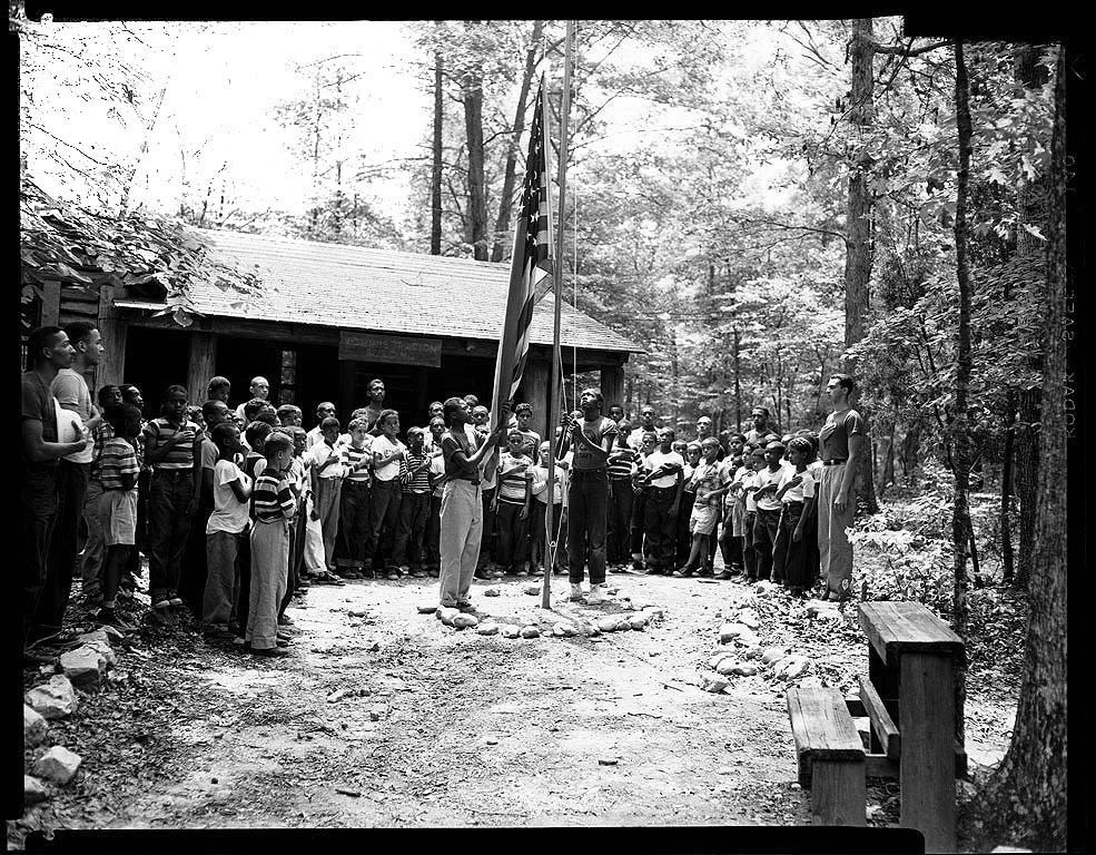 A group of boys of varying heights gather in a semi-circle, hands over hearts, as two boys in in center raise an American flag on a pole.