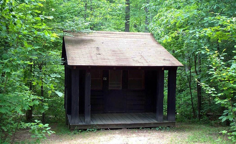 A wooden cabin surrounded by leafy trees has a wooden porch with a roof that is supported by four wooden posts.