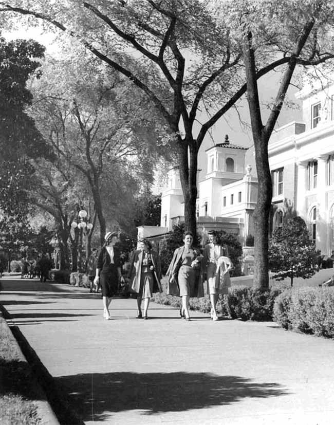 Magnolia Promenade in the late 1940s. Four women walk along a wide promenade past a row of leafy trees and bathhouses.