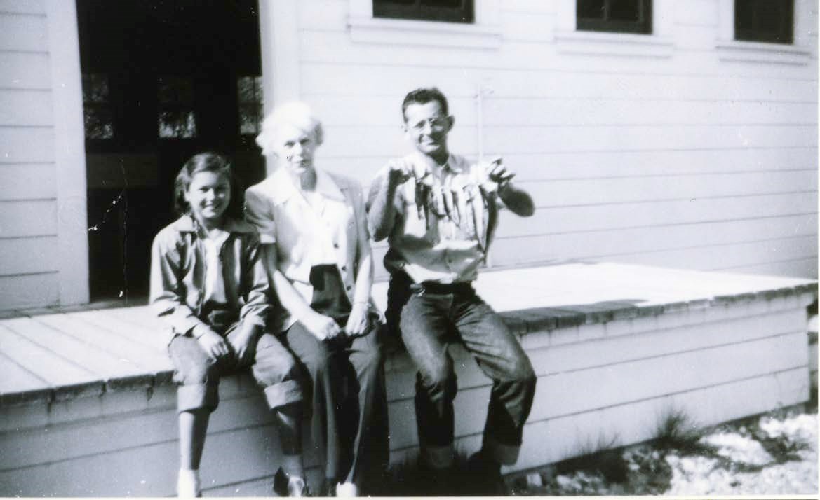 Three people site on a wooden loading dock on a hatchery building. The man is holding fish of varying size on a line.