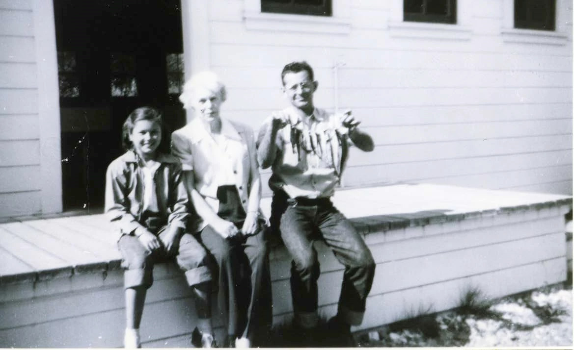 Sitting on the hatchery loading dock, ca. 1943 Three people site on a wooden loading dock on a hatchery building. The man is holding fish of varying size on a line.