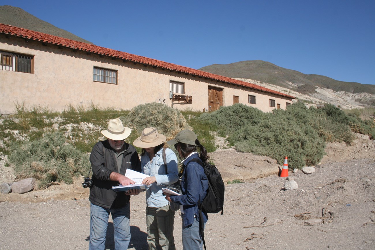 A group of three people with hats and cameras look at a cultural landscape report to assess condition at Death Valley.