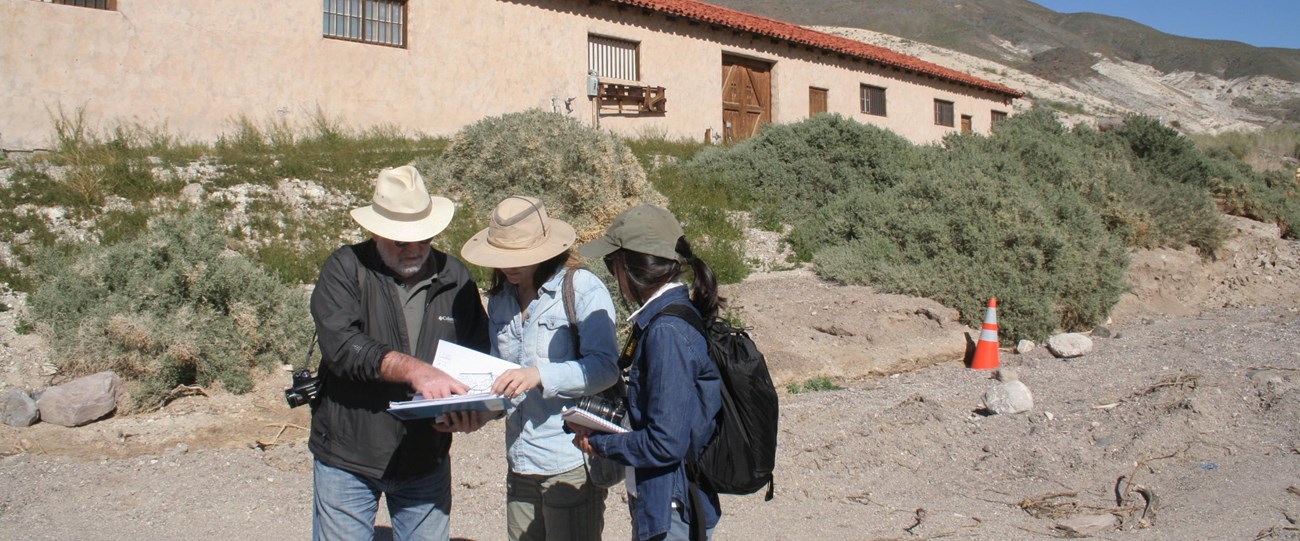 Three people, with cameras and sunhats, look at a notebook at Scotty's Castle, a dry landscape with low vegetation and a long one-story building with terra cotta roof in the background.