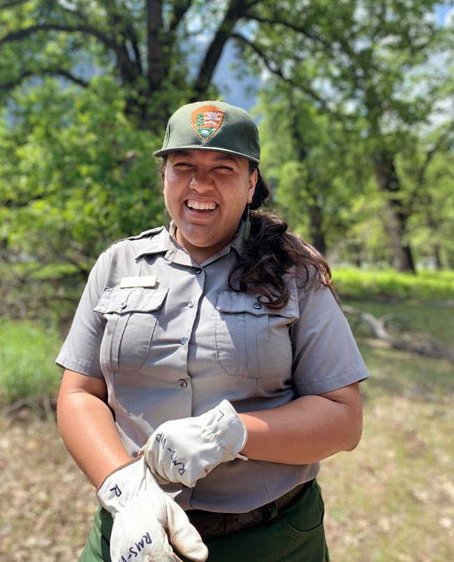 Sandy Hernandez, in NPS uniform, pulls on a pair of work gloves, standing outside in front of trees.