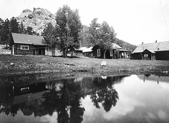 Leafy trees grow beside a grouping of log structures at the edge of a pond, with mountains in the background