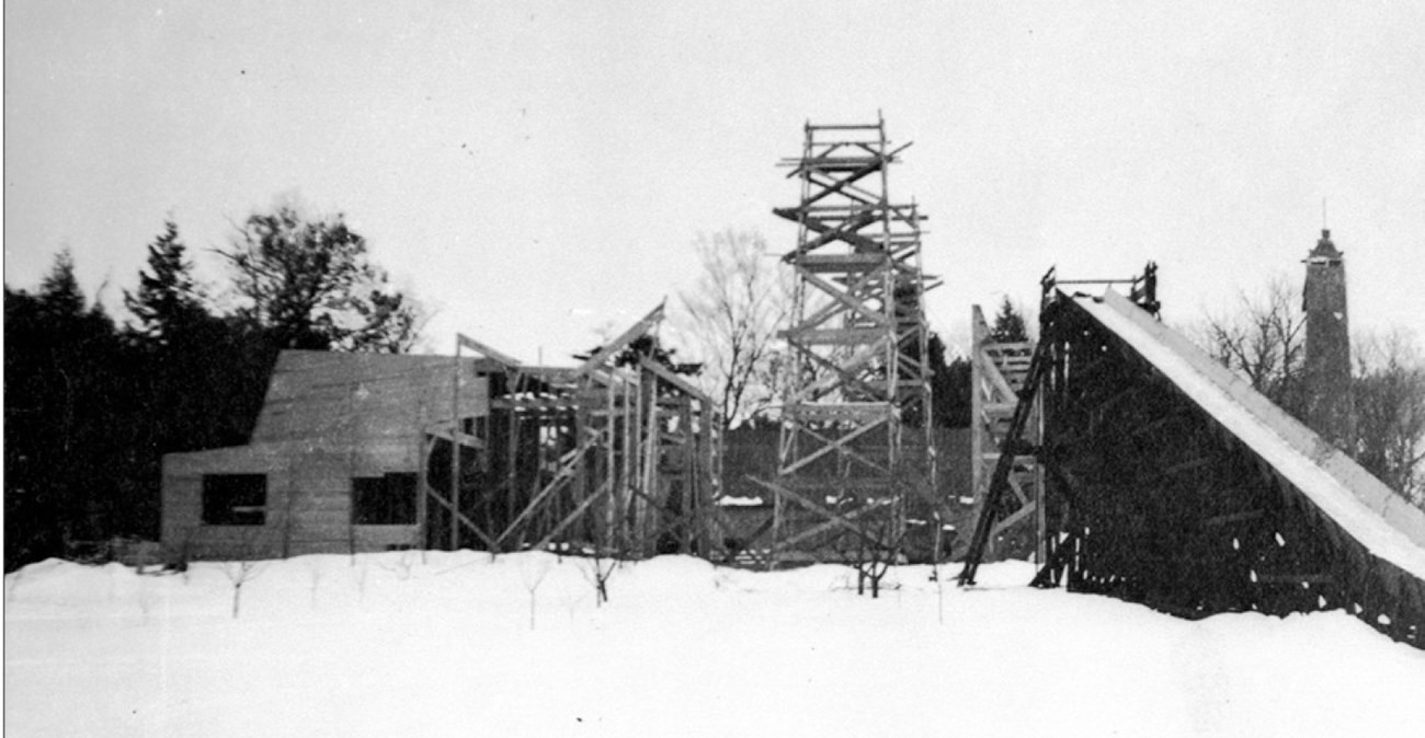 Orchard near the Studio of the Caryatids in 1904, with toboggan run Leafless orchard trees grow in a snowy area in front of a structure under construction. An inclined chute is to the right.