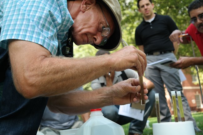 A person kneels and drops something into a test tube of soil and water near three other test tubes, as a group watches.