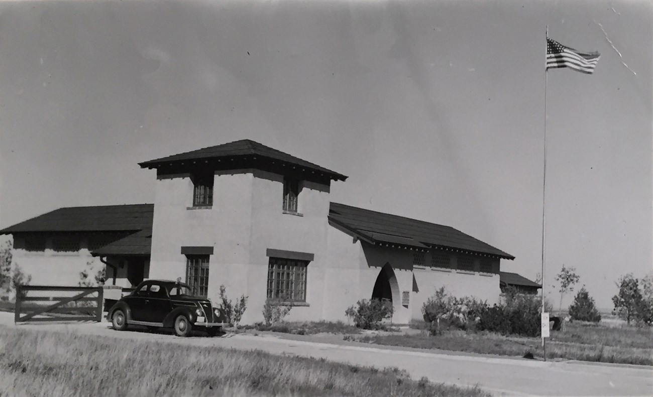 A 1930s style car parked in front of the Scotts Bluff Visitor Center, an adobe structure with a square corner tower. A U.S. flag flies on a pole in front.