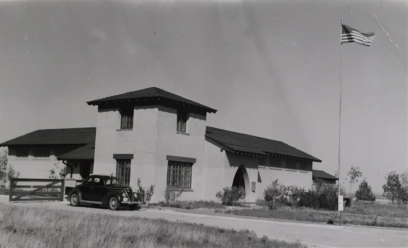 SCBL-Scotts-Bluff-Visitor-Center A 1930s style car parked in front of the Scotts Bluff Visitor Center, an adobe structure with a square corner tower. A U.S. flag flies on a pole in front.