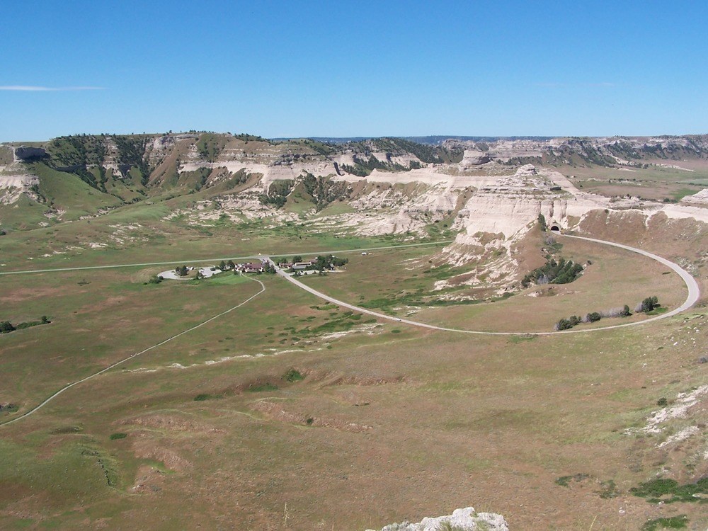 A view of the Scotts Bluff landscape from an overlook includes the Main Complex buildings, rocky bluff, and curving road.