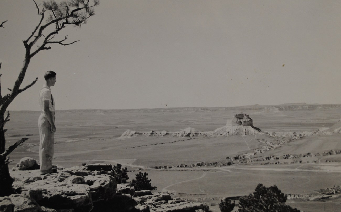 A man stands on a rocky prominence and looks towards Scotts Bluff, a rock formation rising from flat, open plains