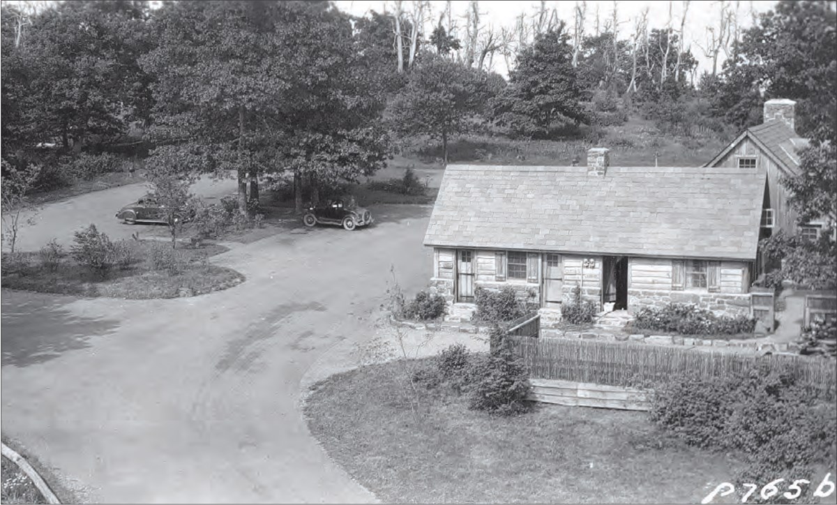 Cars in a parking lot at Wayside station along Skyline Drive in late 1930s. A planted island, CCC installed foundation plantings, and one-story structure surround the lot