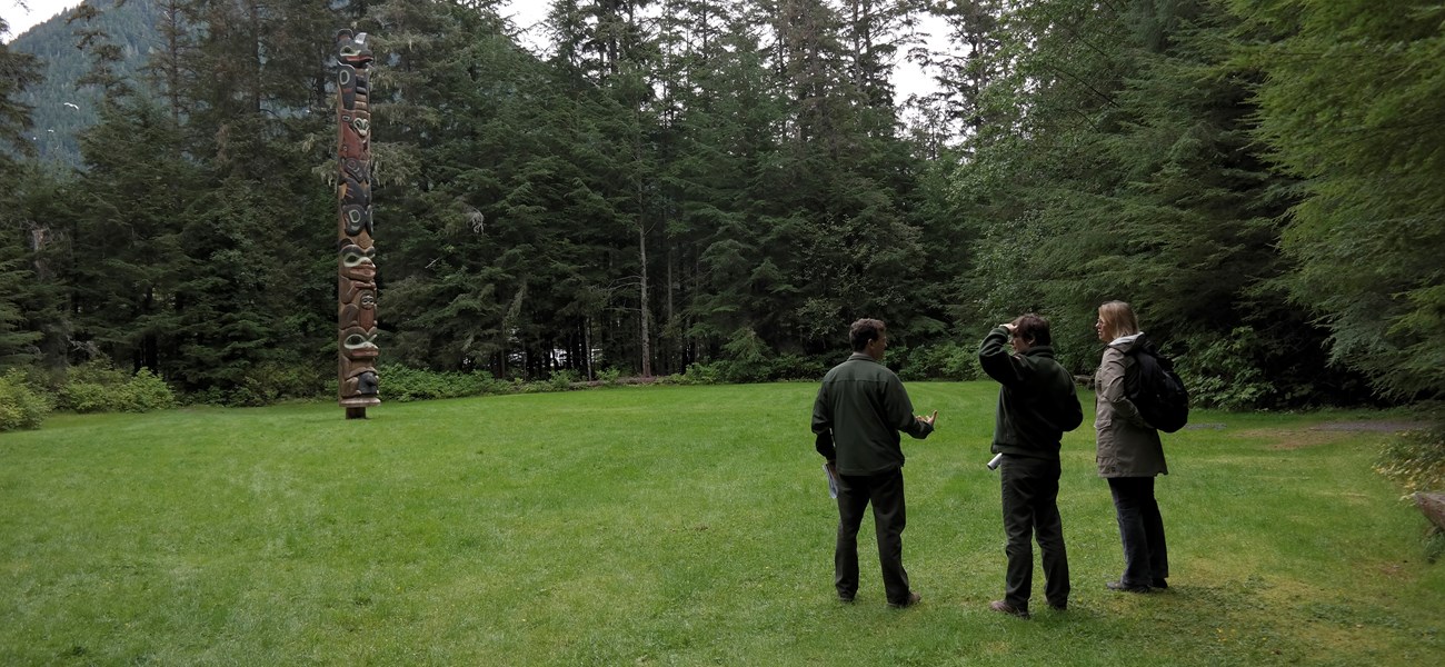 SITK_Sept2014_50 Three people stand in a grassy area facing a totem pole, surrounded by evergreen trees