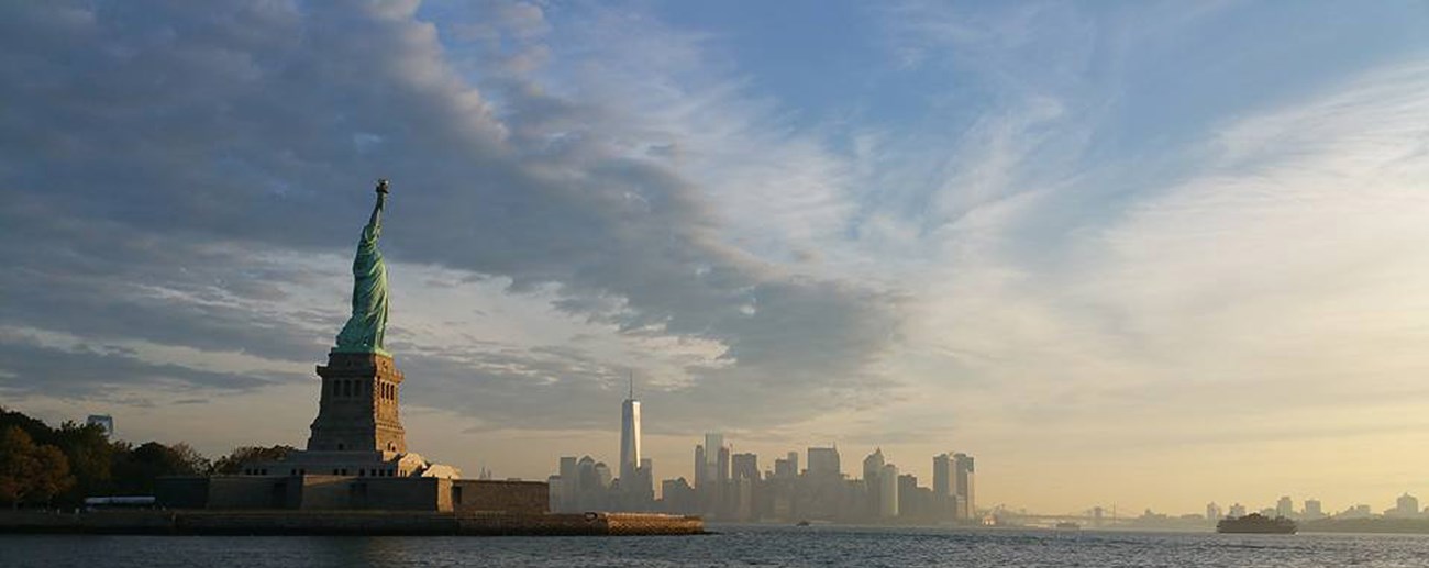 Statue of Liberty on an island in water with the hazy city skyline of New York City in the background.