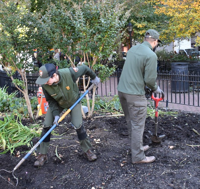 Two employees in NPS uniform work in the soil of a garden, holding a rake and a shovel.