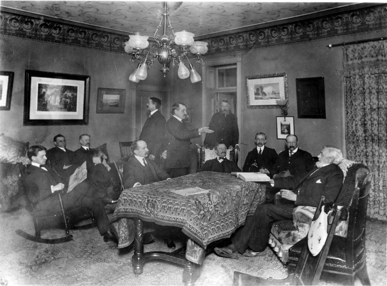 Historic photograph shows a group of well dressed men sitting around an ornate wooden table covered in a fine tablecloth in a richly furnished room.