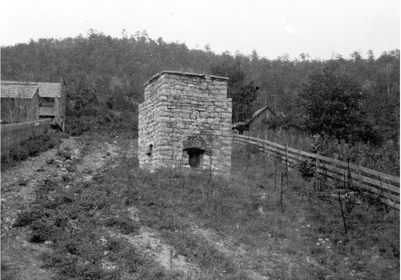 Smelter-historic-BUFF Rush HD CLR EA 3_2019 196 Rectangular brick Rush Smelter with an oven-like opening in the side. A road to the left leads to wooden buildings.