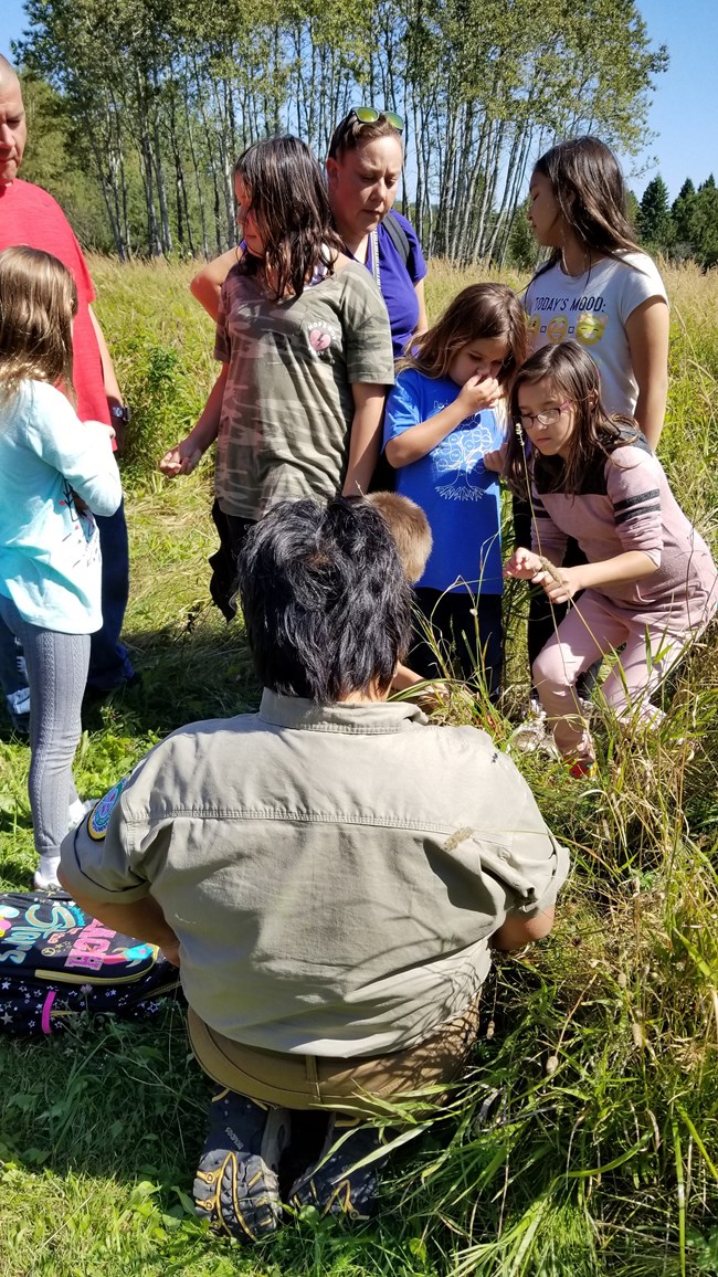 Young students gather in front of someone kneeling in a grassy area