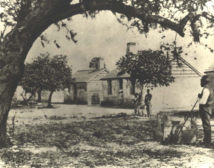 Historic image of an African American man standing beside a square well under a tree, near a row of small symmetrical cabins.