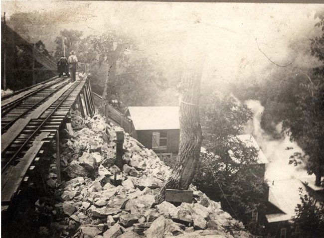 A person walks on an elevated tram way, over a rocky hillside towards mill structures on the hillside.