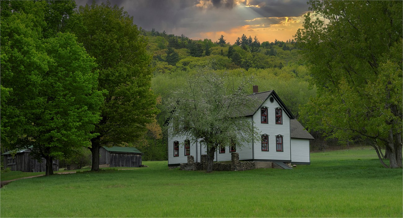 A cloudy sky glows over a farm, with a two-story farm house, tall apple tree, and outbuildings
