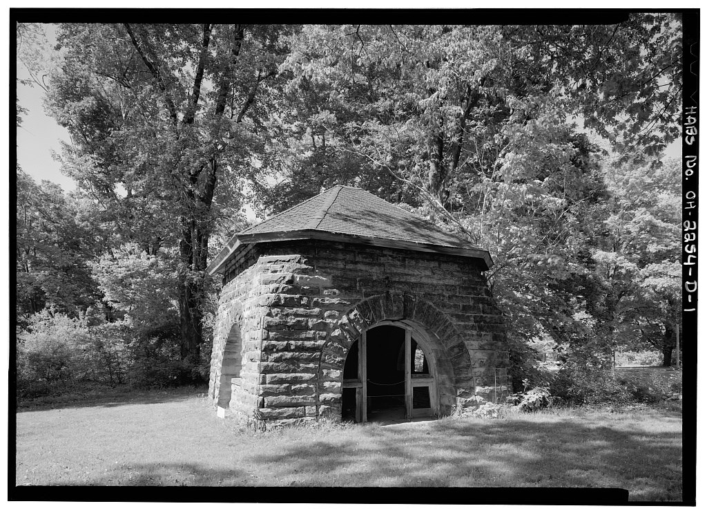 A rectangular stone structure with arched open doorways and shingle roof, framed by trees
