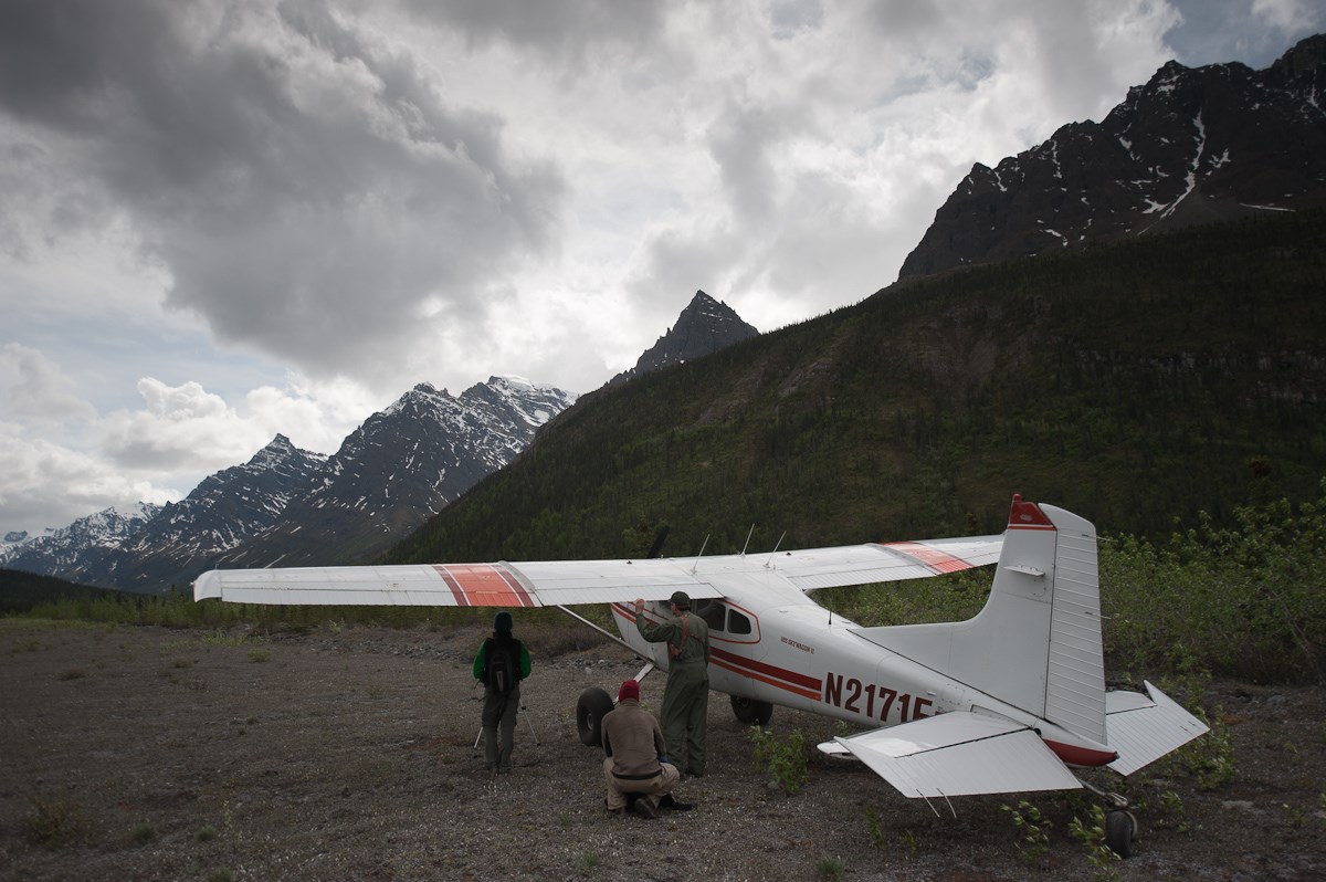 Three people stand under the wing of a small plane on a backcountry airstrip, surrounded by mountains.