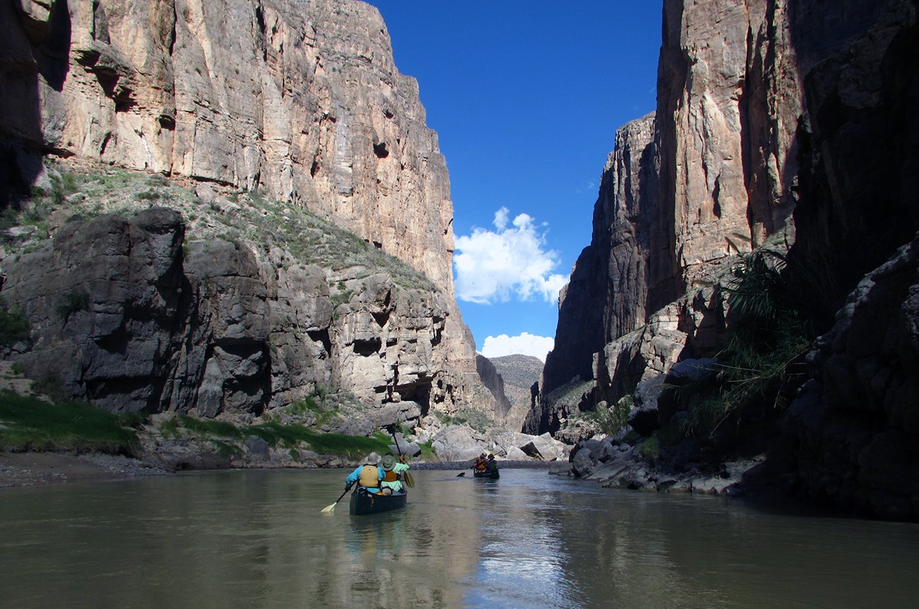 Two canoes float down a smooth river between two tall cliff walls