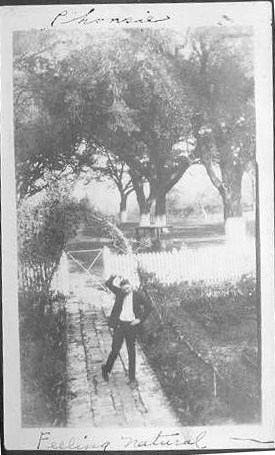 A grainy photograph of a man standing on a garden path, with a white picket fense and tall trees in the background