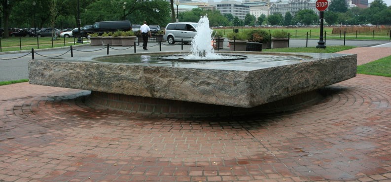 Water sprays up out of the center of fountain that is round pool of water in a stone square base, surrounded by a red brick patio and an urban landscape