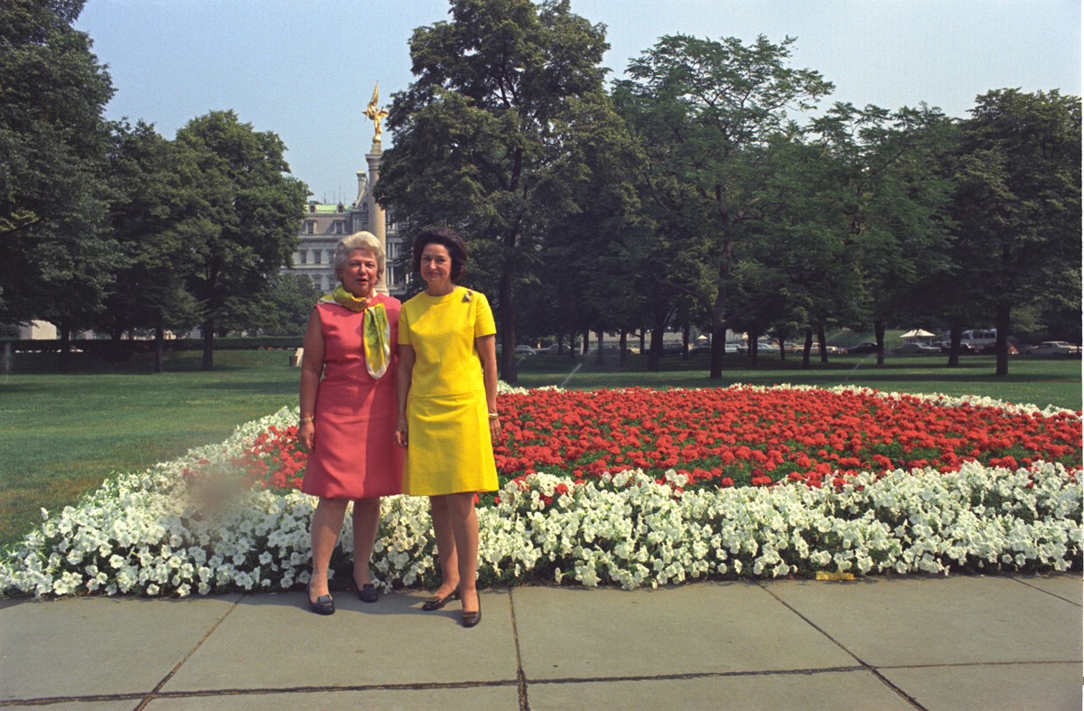 Two women in brightly-colored dresses pose on a sidewalk in front of a flower bed, with white-blooming flowers forming a border around red blooming flowers.