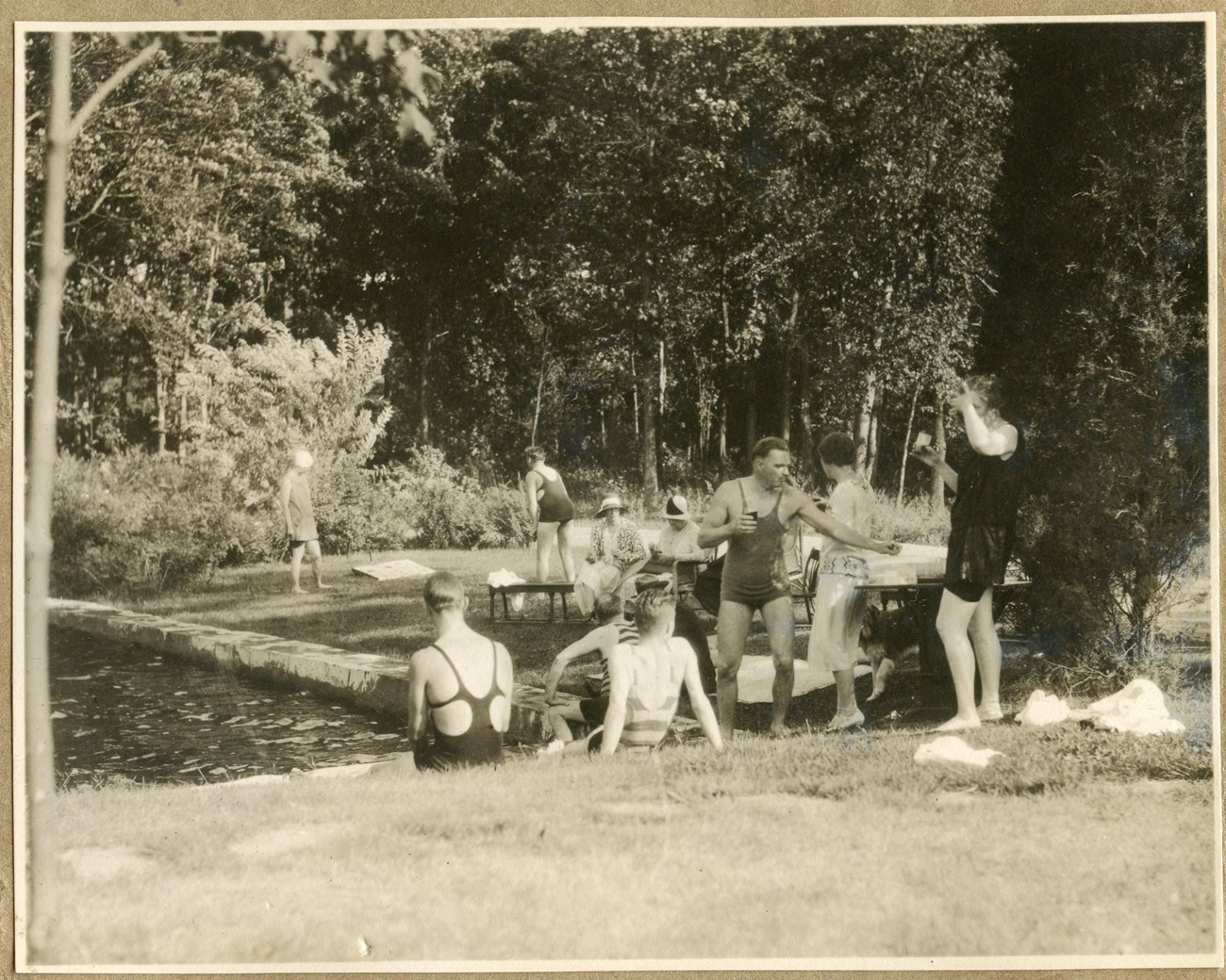 A group of people wearing 1930s style swimwear gather in the grass at the edge of a pool
