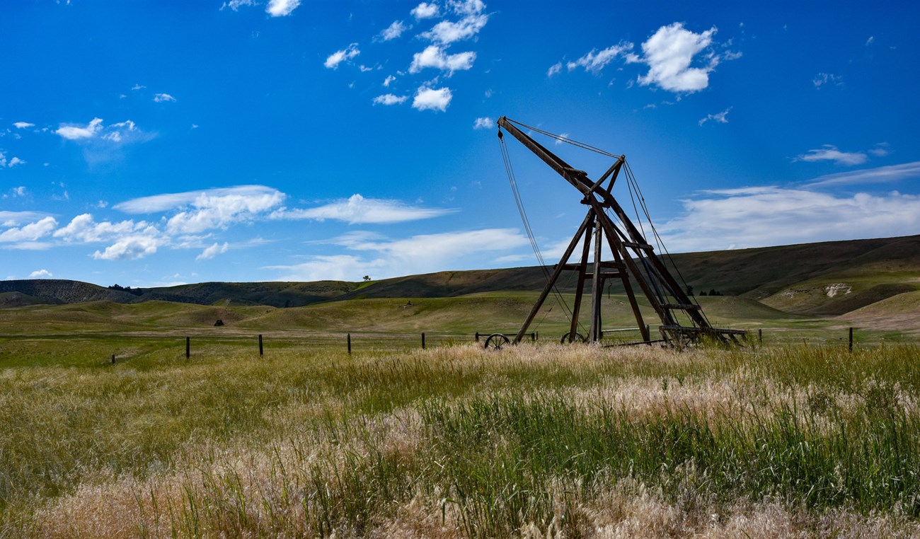 Hay Rake Open, green hay fields and distant rolling hills. A large piece of equipment stands in the center, a collection of ropes, pulleys, and framework that allow for the collection of hay into bales